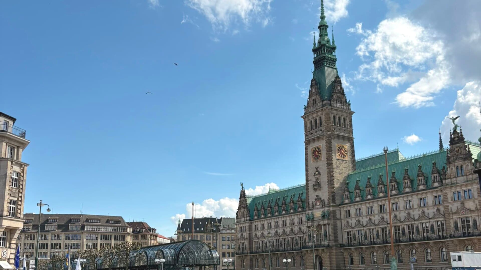 Das Hamburger Rathaus mit grünem Kupferdach und Turm am Rathausmarkt, darunter der gläserne Alsterarkaden-Pavillon, an einem sonnigen Tag mit blauem Himmel